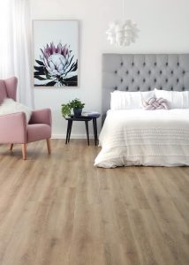 Bedroom scene featuring luxurious bed, soft pink chair, side table, plant wall art and floors by Imagine Floors by Airstep
